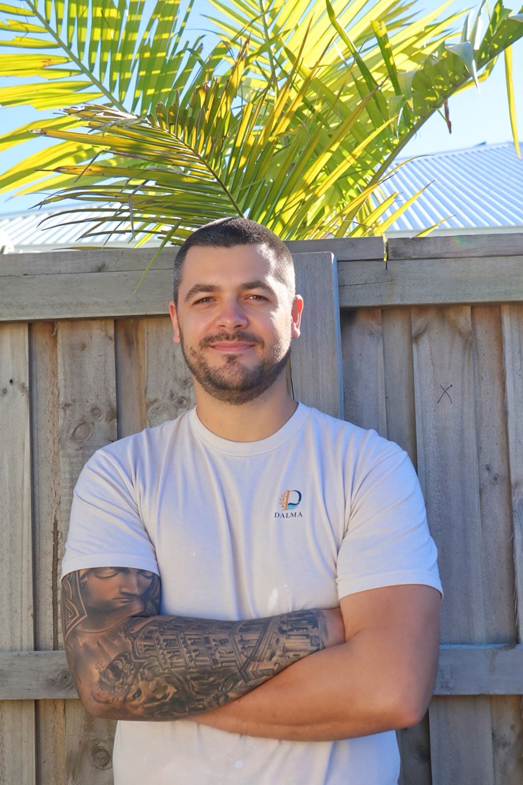Bearded man with arm tattoos standing before a wooden fence and palm tree.