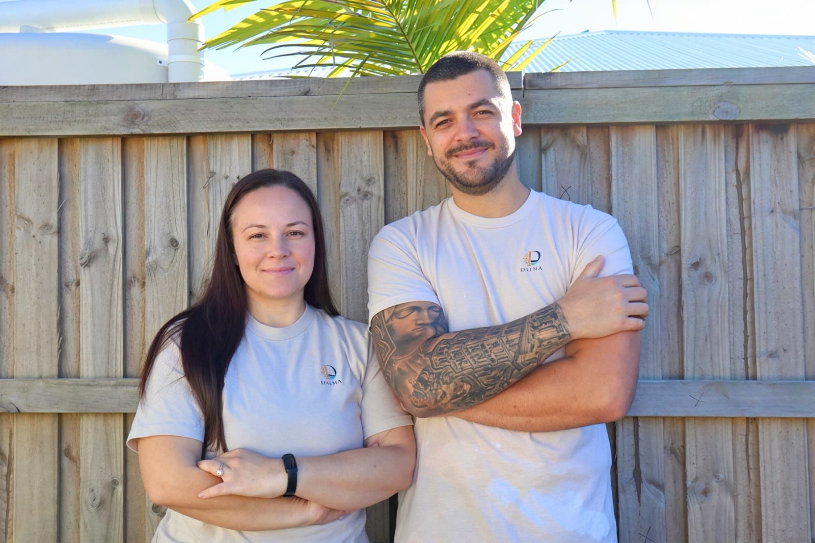 A man and woman standing with arms crossed in front of a wooden fence outdoors.