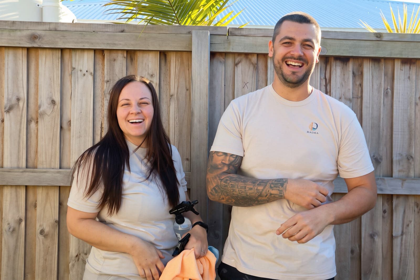 A man and woman laughing in front of a wooden fence, holding a spray bottle.