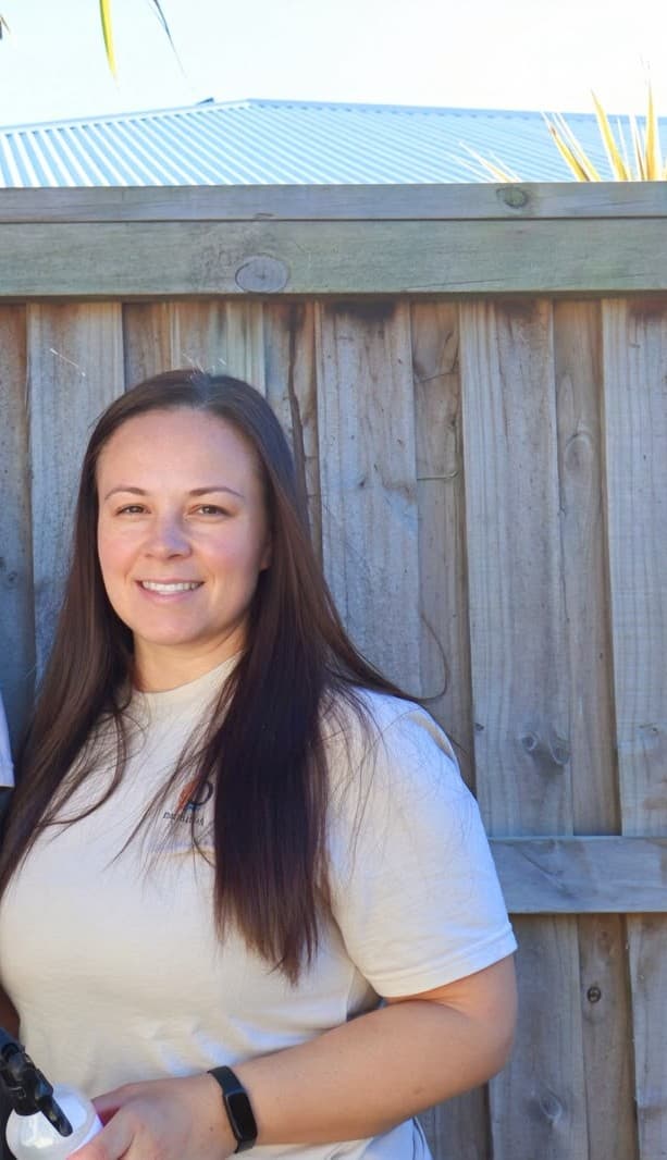 Smiling woman with long brown hair wearing a white shirt against a wooden fence.